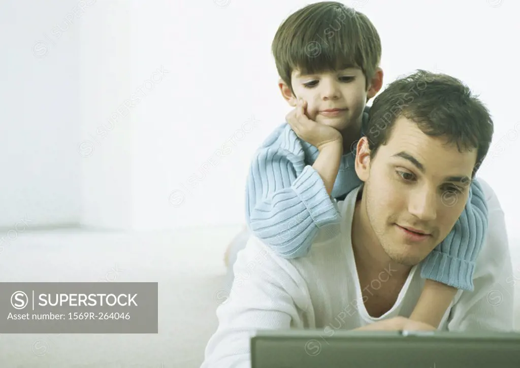 Little boy on man´s back, both looking down at laptop