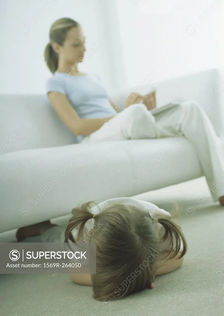 Little girl lying face down on floor, young woman sitting on sofa, reading
