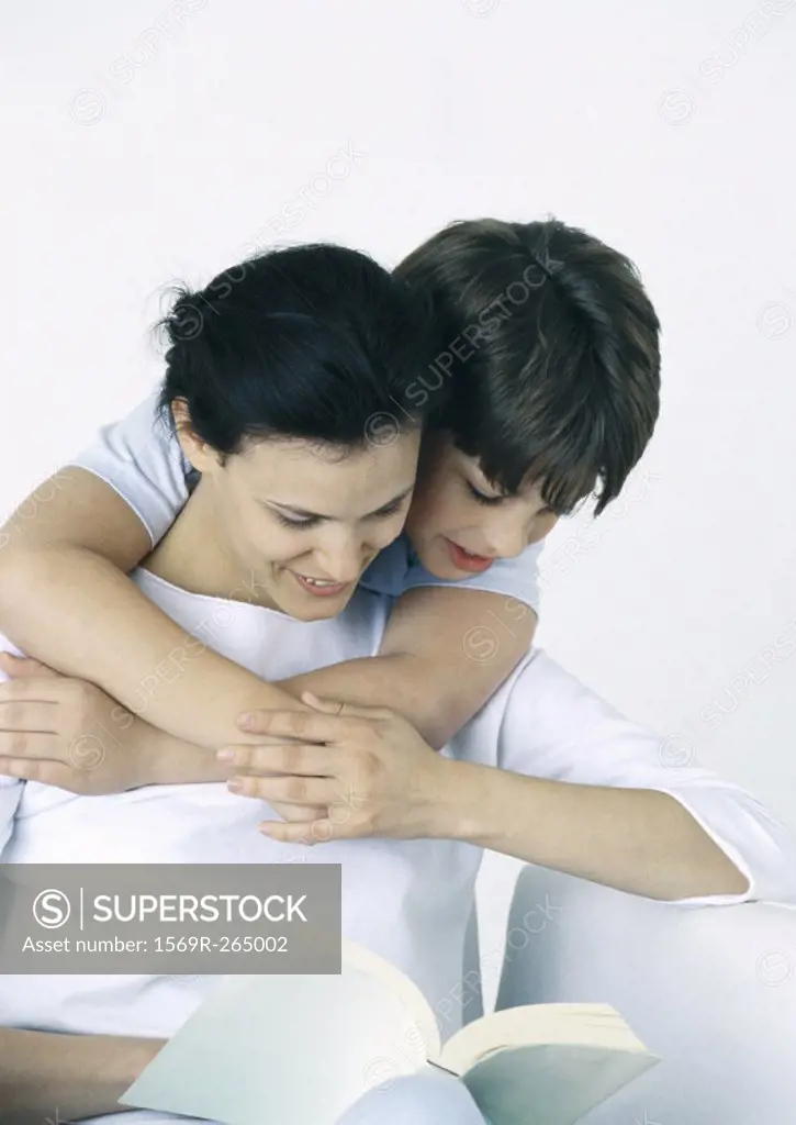 Woman and son reading book, boy with arms around woman, looking over her shoulder
