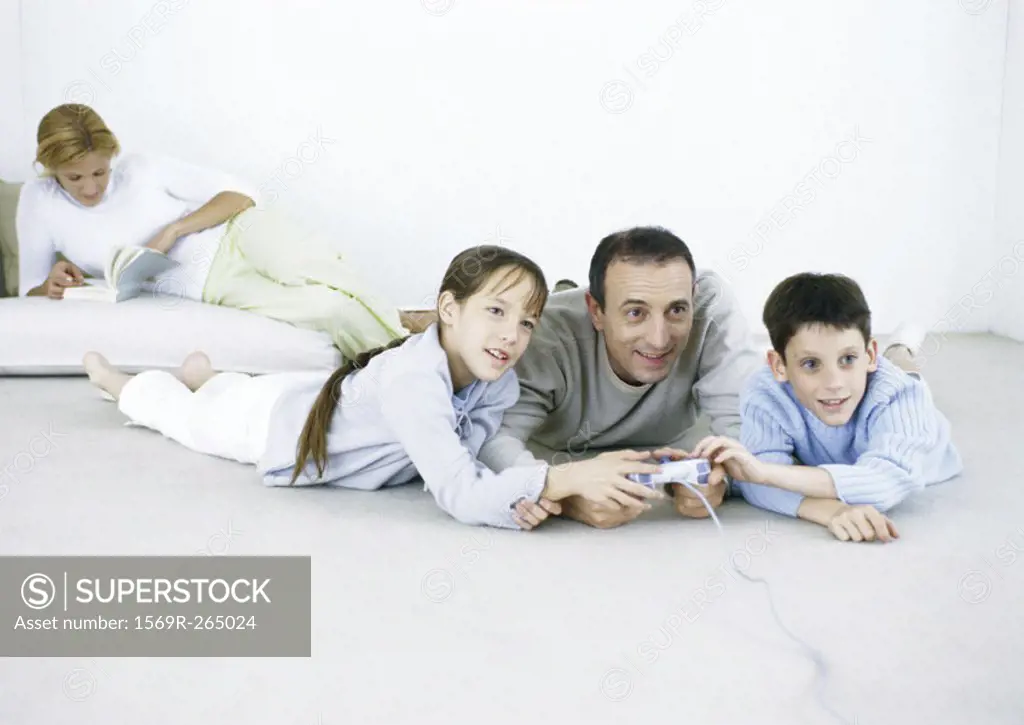 Man and boy and girl lying on floor playing video game, woman reading in background