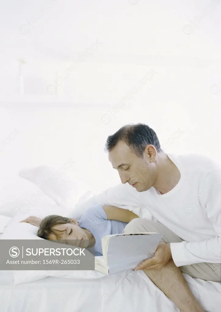 Man sitting on edge of bed reading book to girl lying in bed