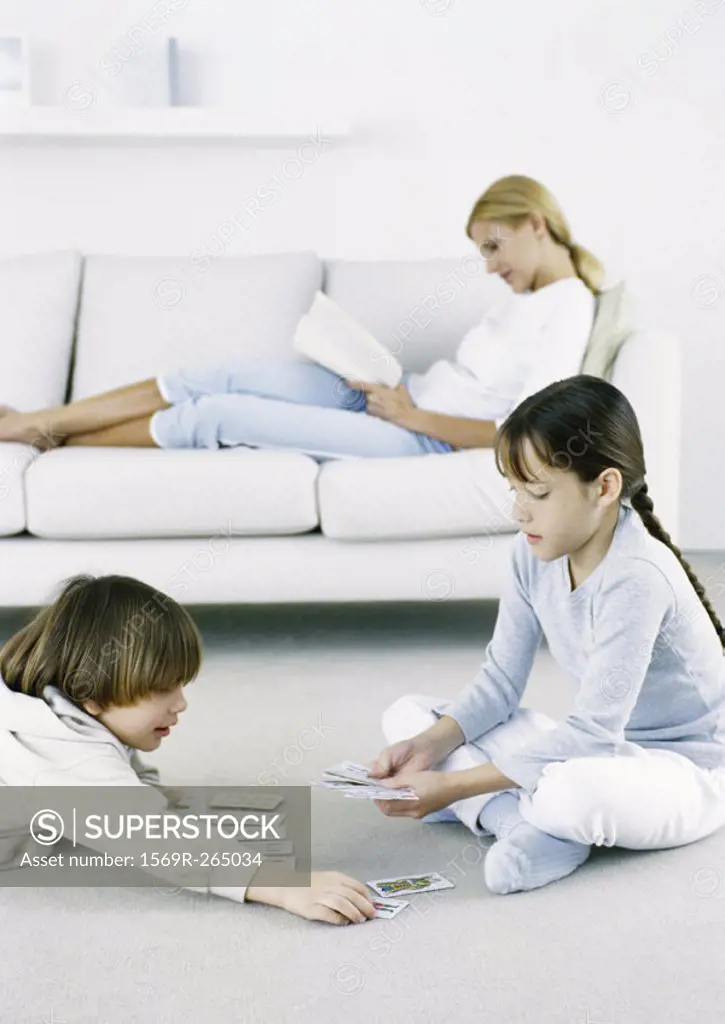 Girl and boy playing cards on floor, woman sitting on sofa reading in background