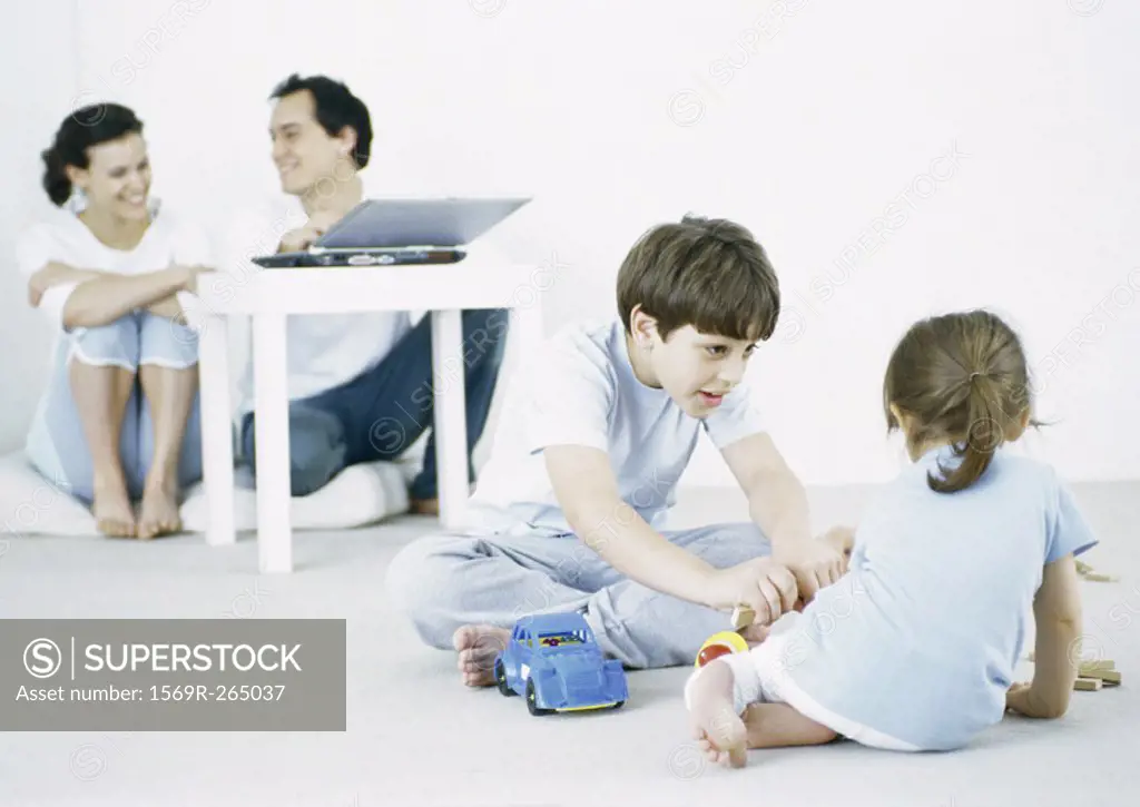 Boy and girl playing on floor, parents using laptop in background