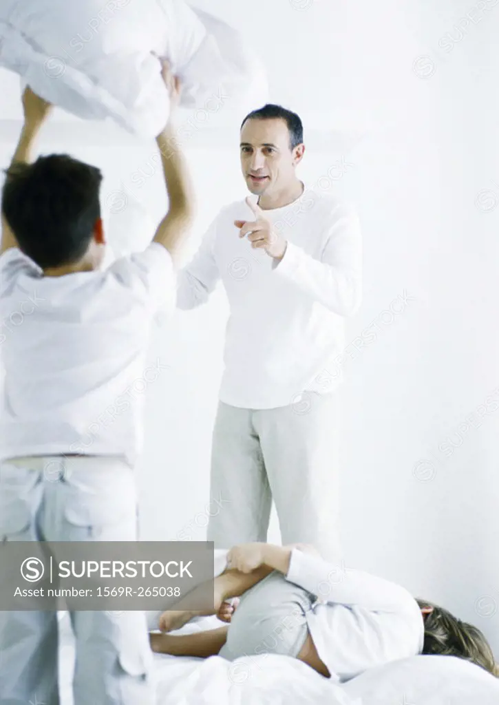Boy standing on bed holding up pillow, rear view, second boy lying on bed, man pointing at first boy