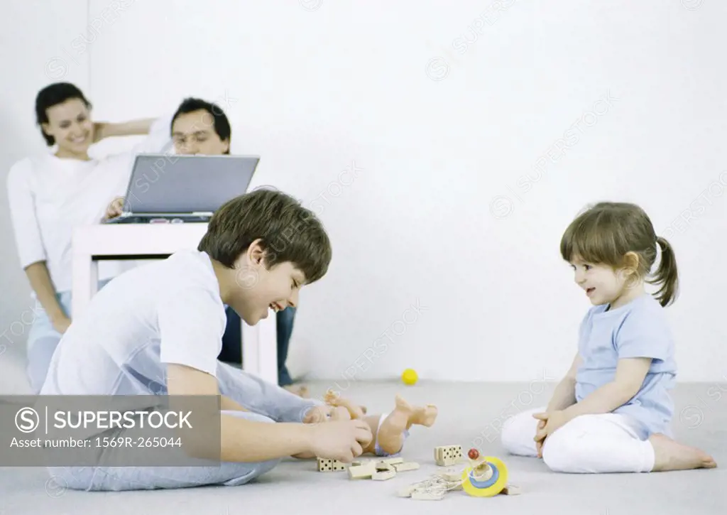 Boy and girl sitting on floor playing, man and woman using laptop in background