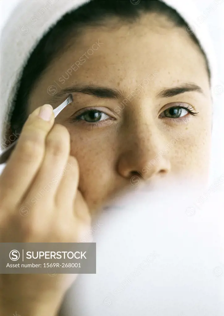 Young woman tweezing eyebrows, compact blurred in foreground