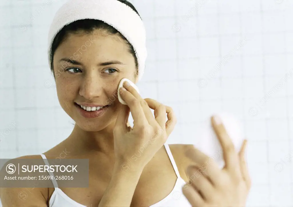 Woman wiping face with cotton pad, looking at compact