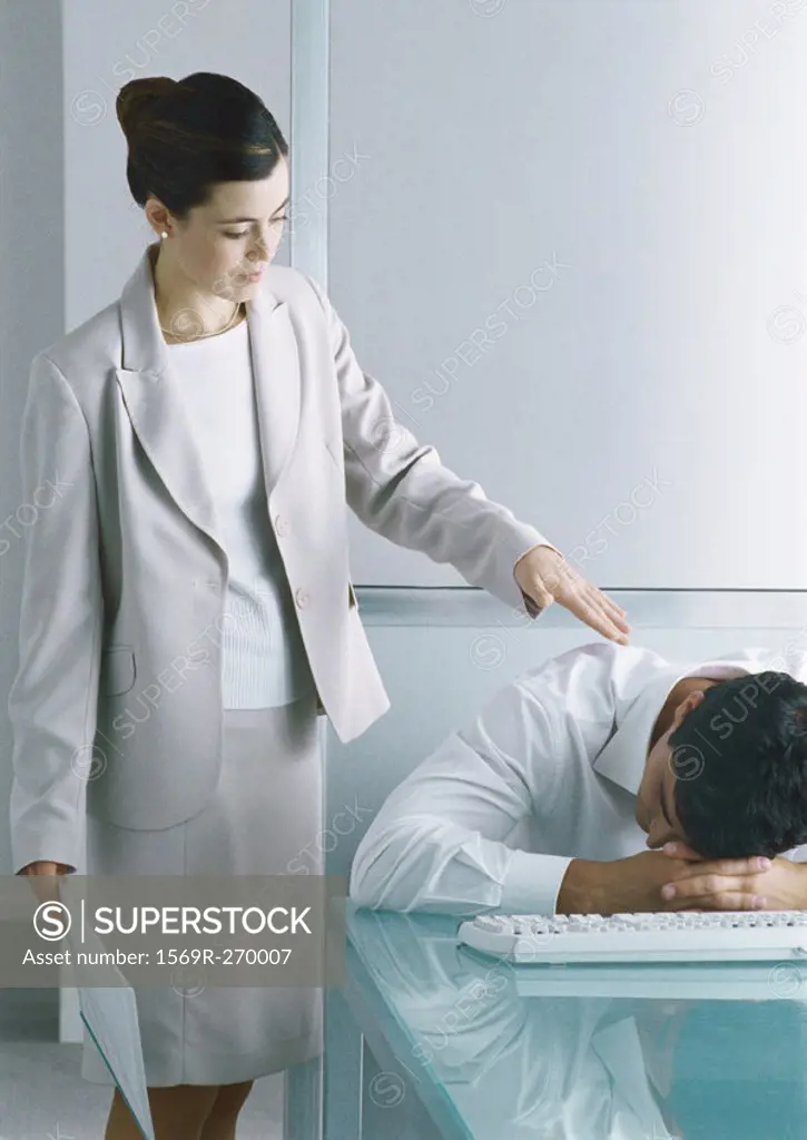 Man sitting with head down on desk, woman standing next to him tapping his shoulder