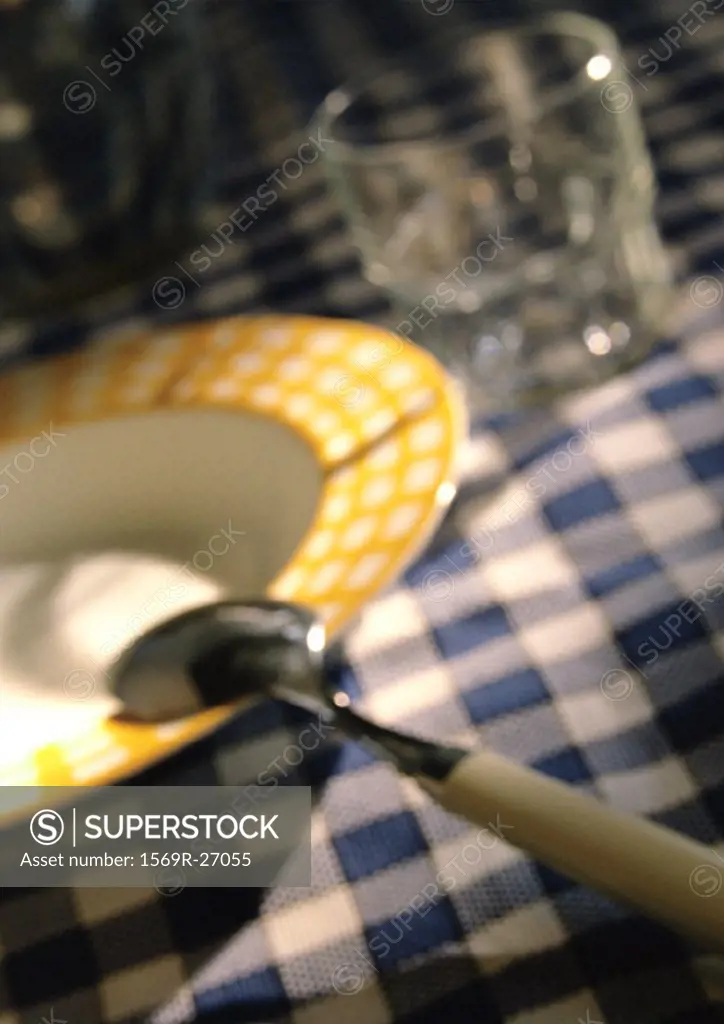 Bowl, spoon and glass on checked tablecloth, close-up, blurred