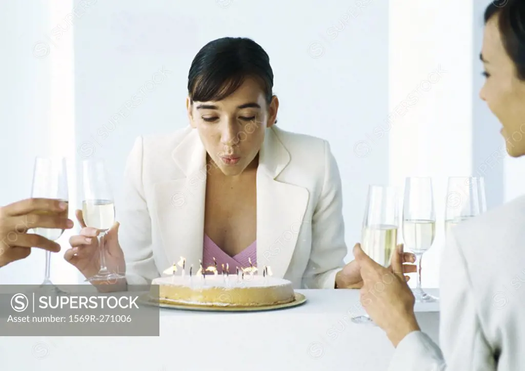 Woman blowing out candles on cake, sitting with other people, holding glasses of champagne