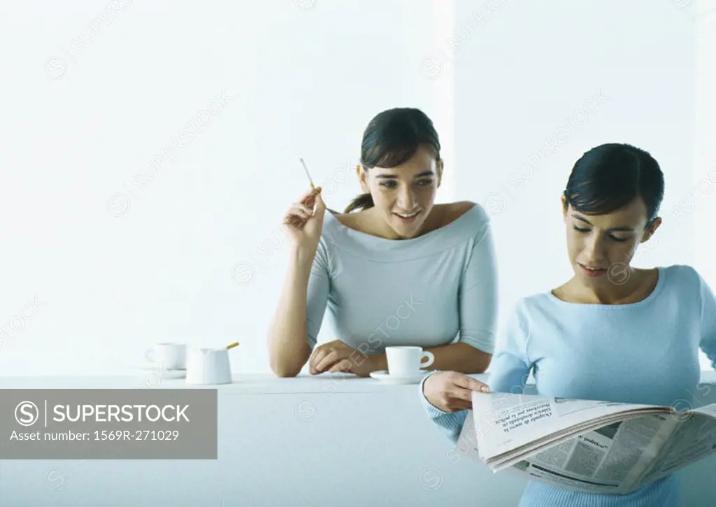 Two women taking coffee break, one reading newspaper, one smoking