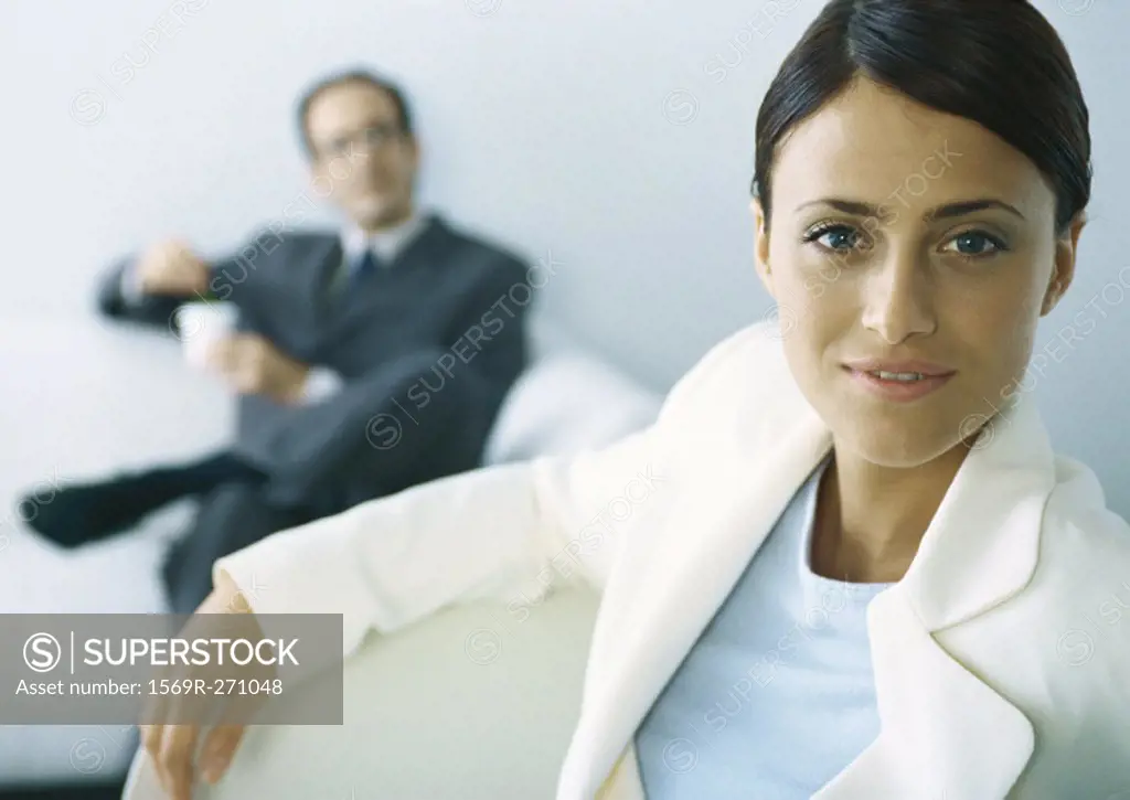 Businesswoman looking at camera, businessman with legs crosses holding cup in background