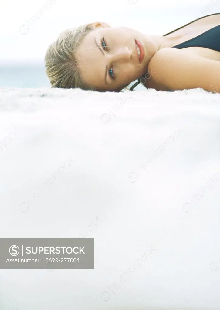 Young woman in bathing suit lying on sand, looking at camera, head and shoulders