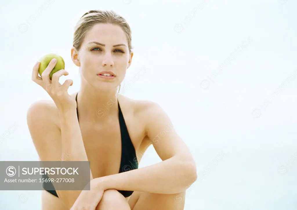 Young woman in bikini sitting with knees up and holding apple