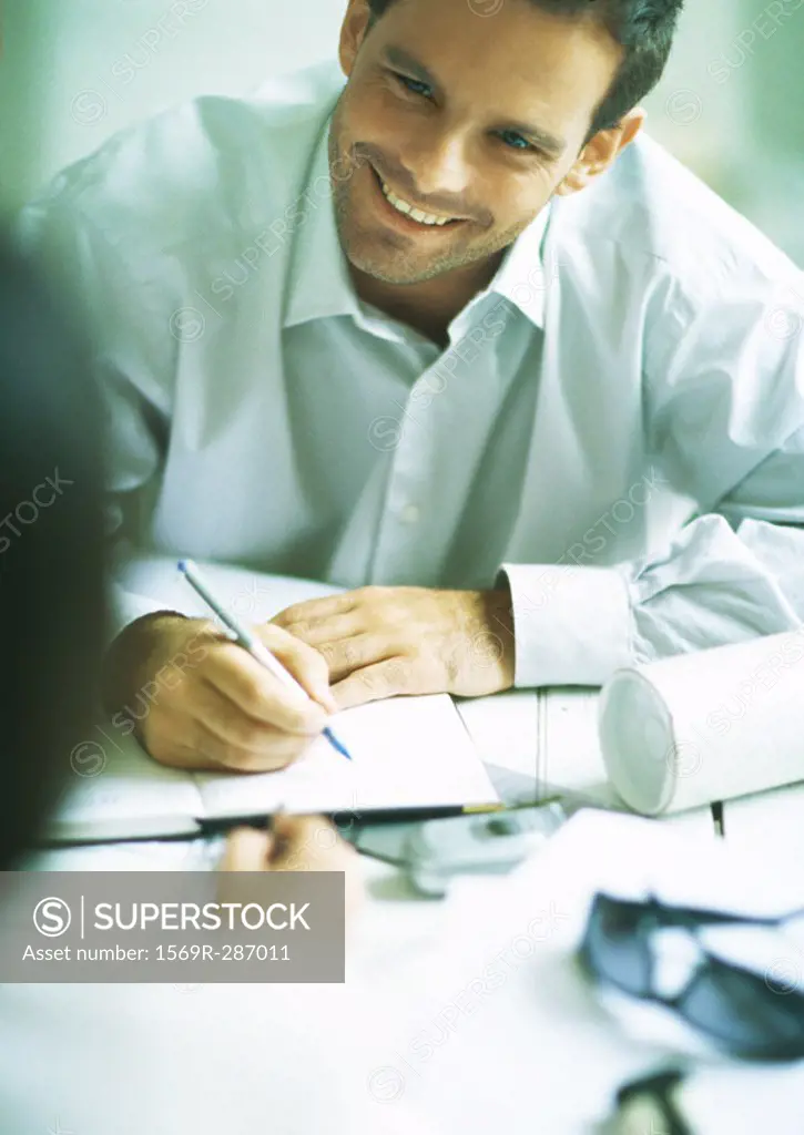 Man smiling and writing in agenda, looking at second person