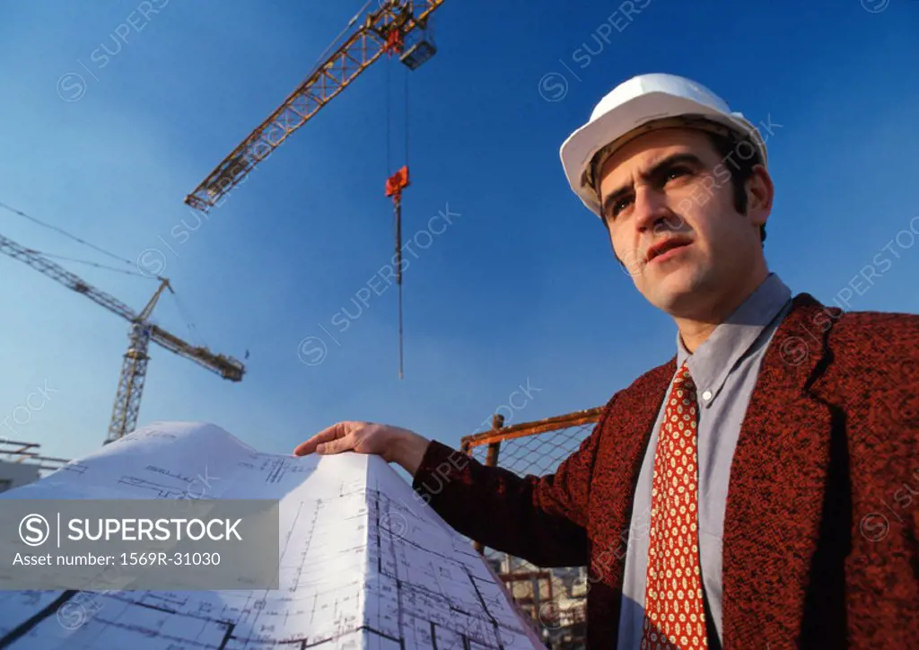 Man wearing hard hat, holding blueprint, cranes in background, low angle view