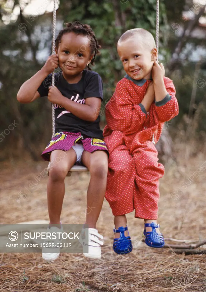 Little boy and girl sitting on swing together, each holding one side of swing rope, smiling at camera, full length