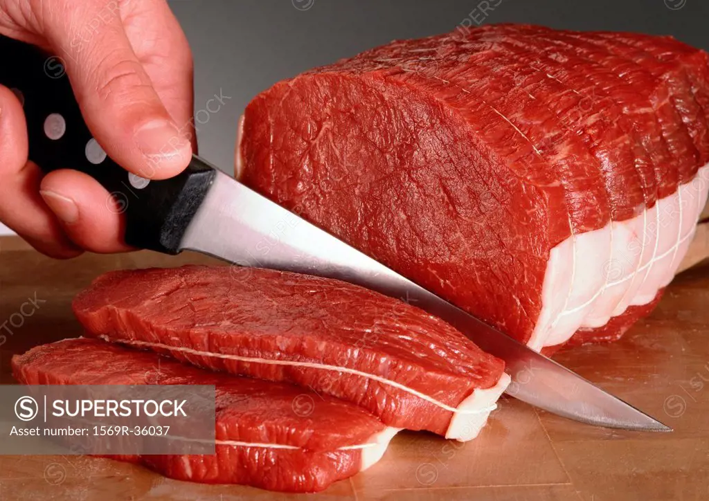Fingers holding knife, slicing raw beef fillets, close-up
