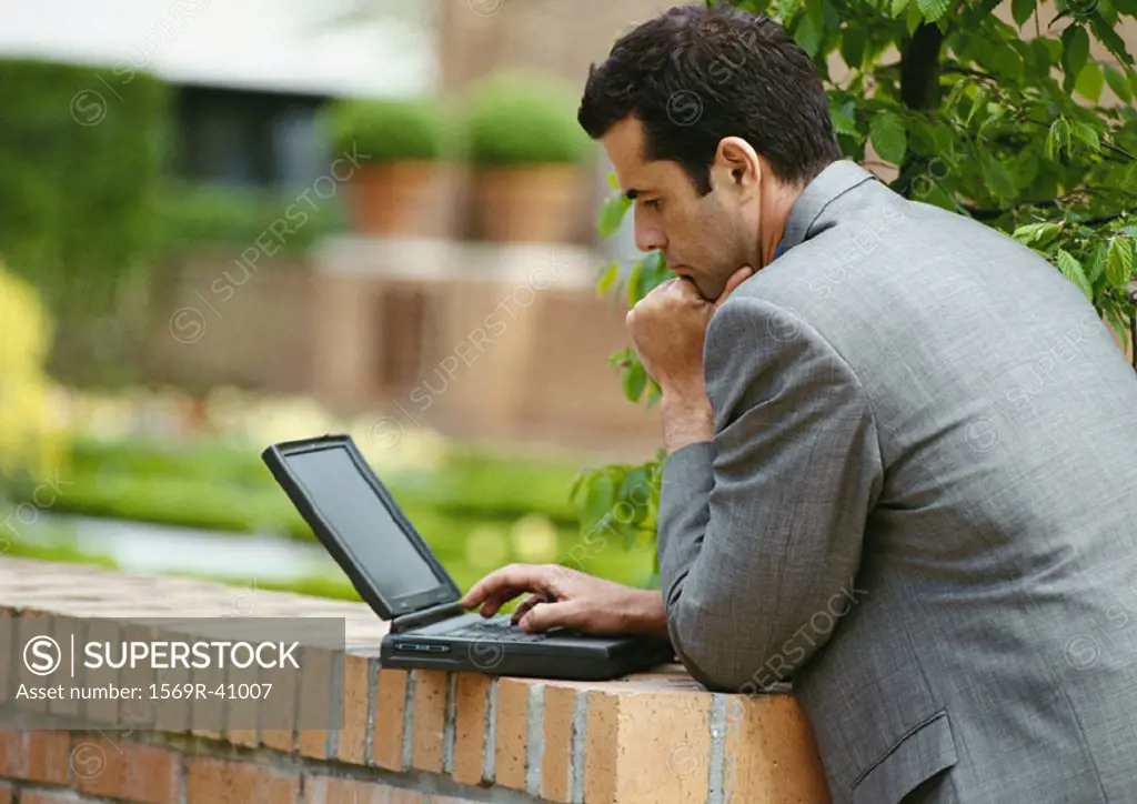 Businessman using laptop computer outside, leaning against low brick wall, side view