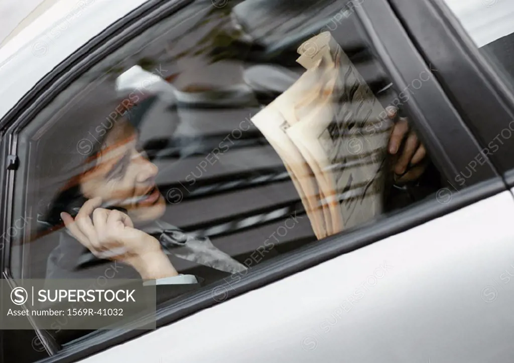 Businessman sitting in back of car, holding newspaper and cell phone, viewed through window