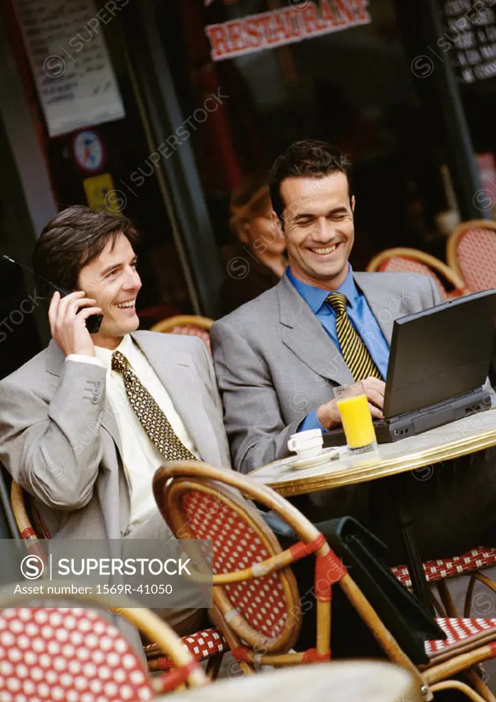 Two businessmen sitting at cafe terrace, using cell phone and laptop computer