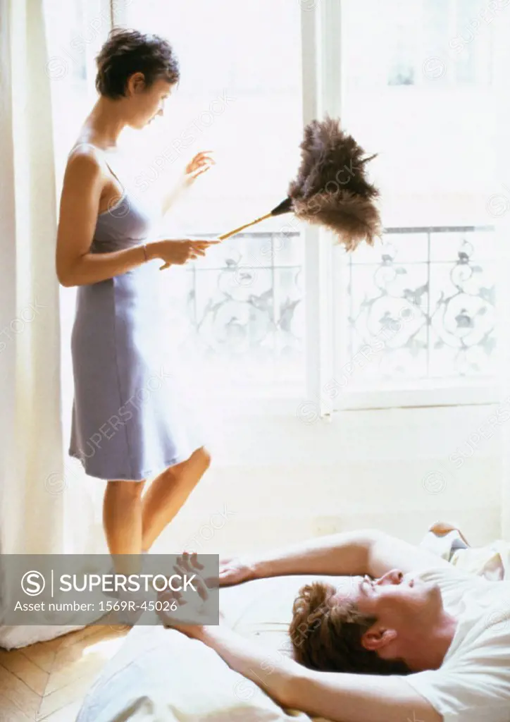 Woman holding feather duster by window, full length, man lying on bed