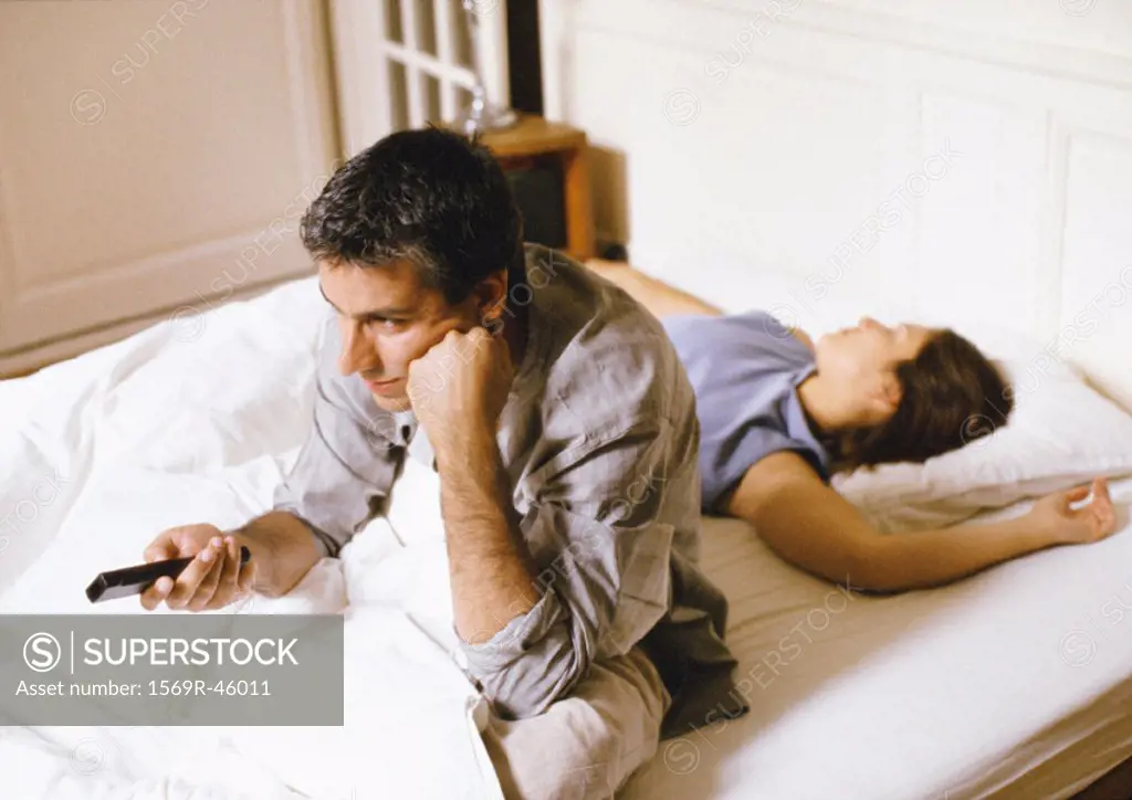 Man sitting up in bed, pointing remote control, woman lying behind him