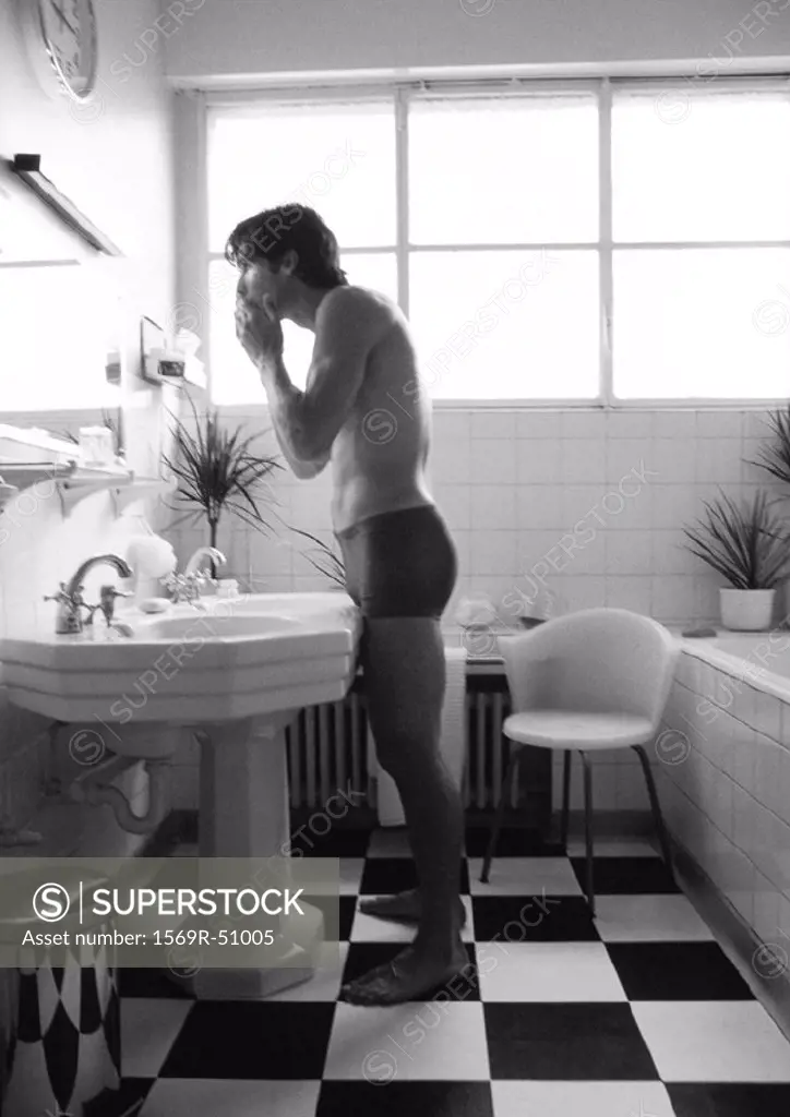 Man in underwear standing, looking in mirror in bathroom, side view, b&w
