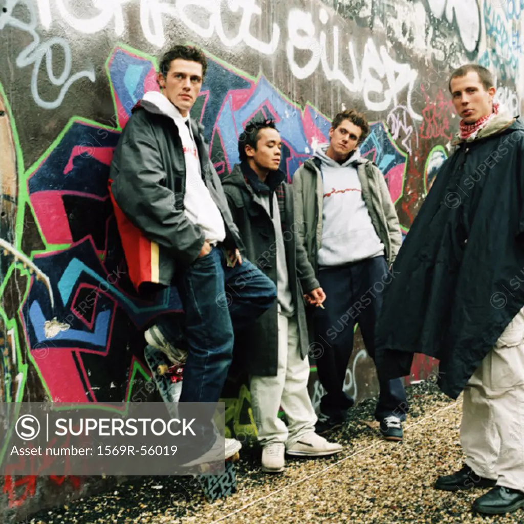 Four young men in city street, three leaning against graffiti wall