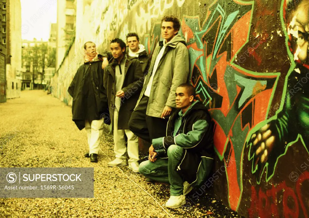 Five young men leaning against wall covered in graffiti, full length