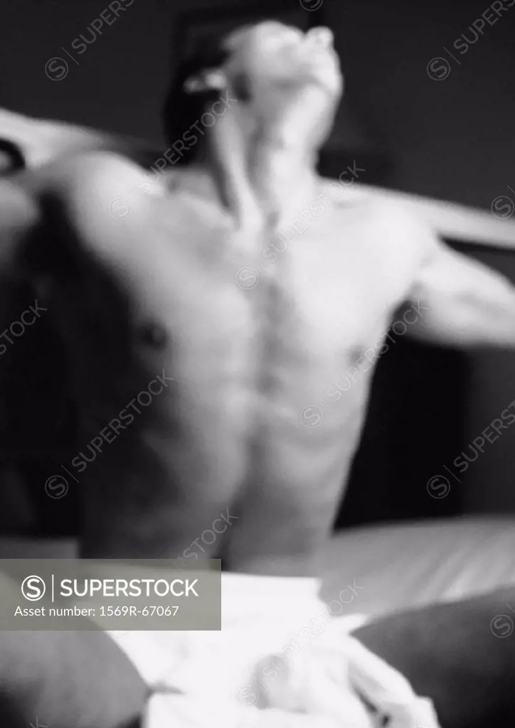 Man sitting on bed in underwear, leaning head back and stretching arms out wide, blurred black and white