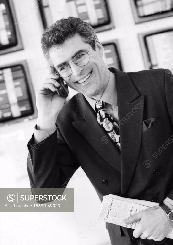 Businessman using cellular phone in airport, smiling and holding newspaper in front of monitors, black and white portrait