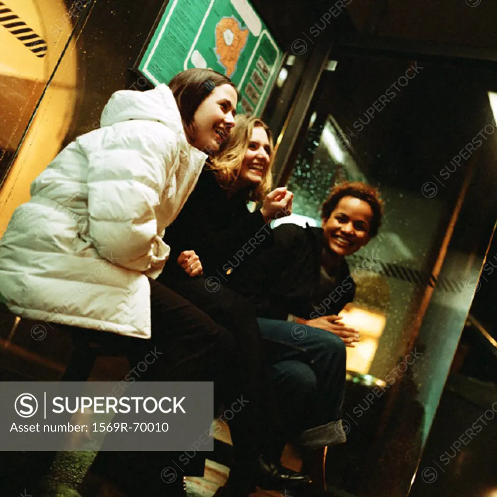 Three young women sitting in bus shelter at night, laughing