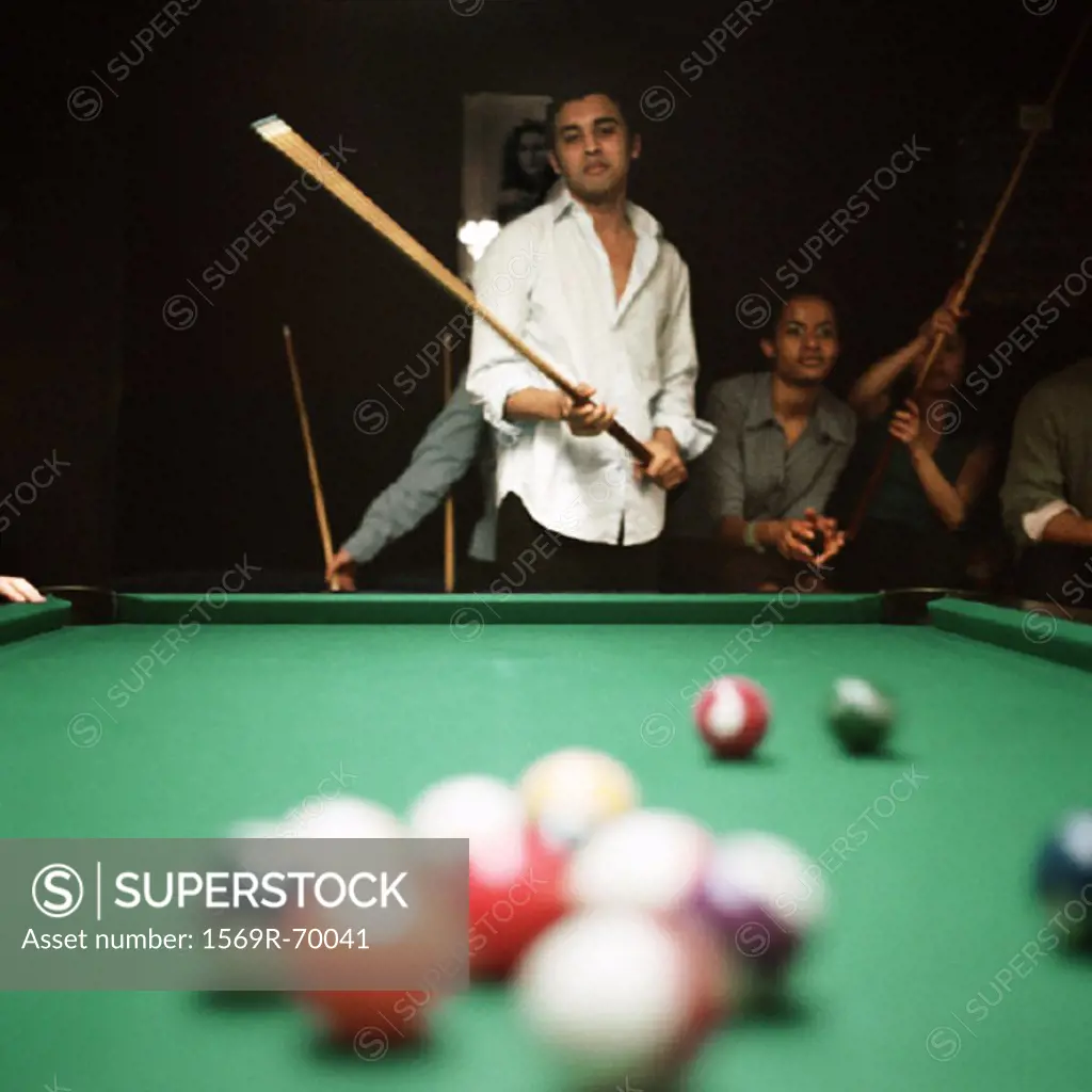 Young man shooting pool, people watching in background, billiard balls blurred in foreground