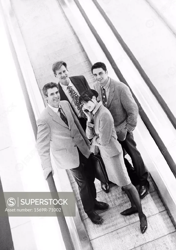 Group of business people on moving walkway smiling at camera, business woman in front on cell phone, b&w