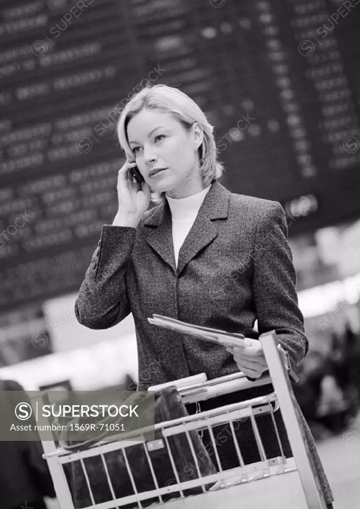 Businesswoman on cell phone with luggage caddy, in front of arrival and departure board, b&w