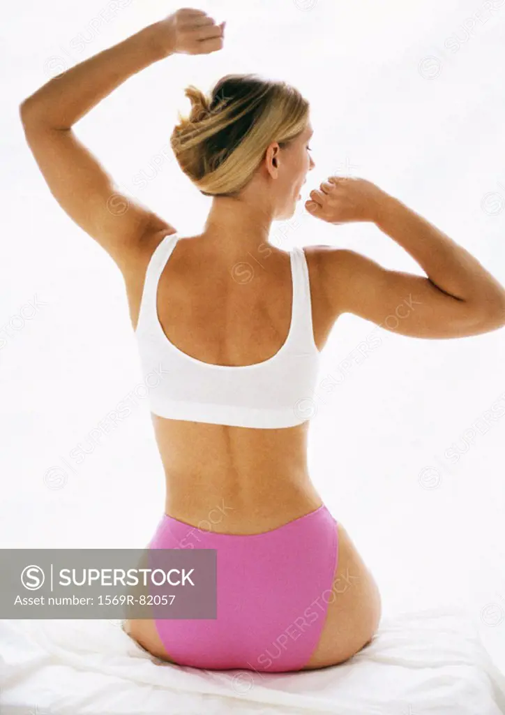 Woman sitting in underwear, stretching arms, rear view