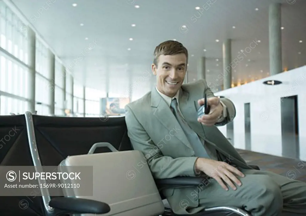 Businessman sitting in airport lounge, holding out cell phone toward camera