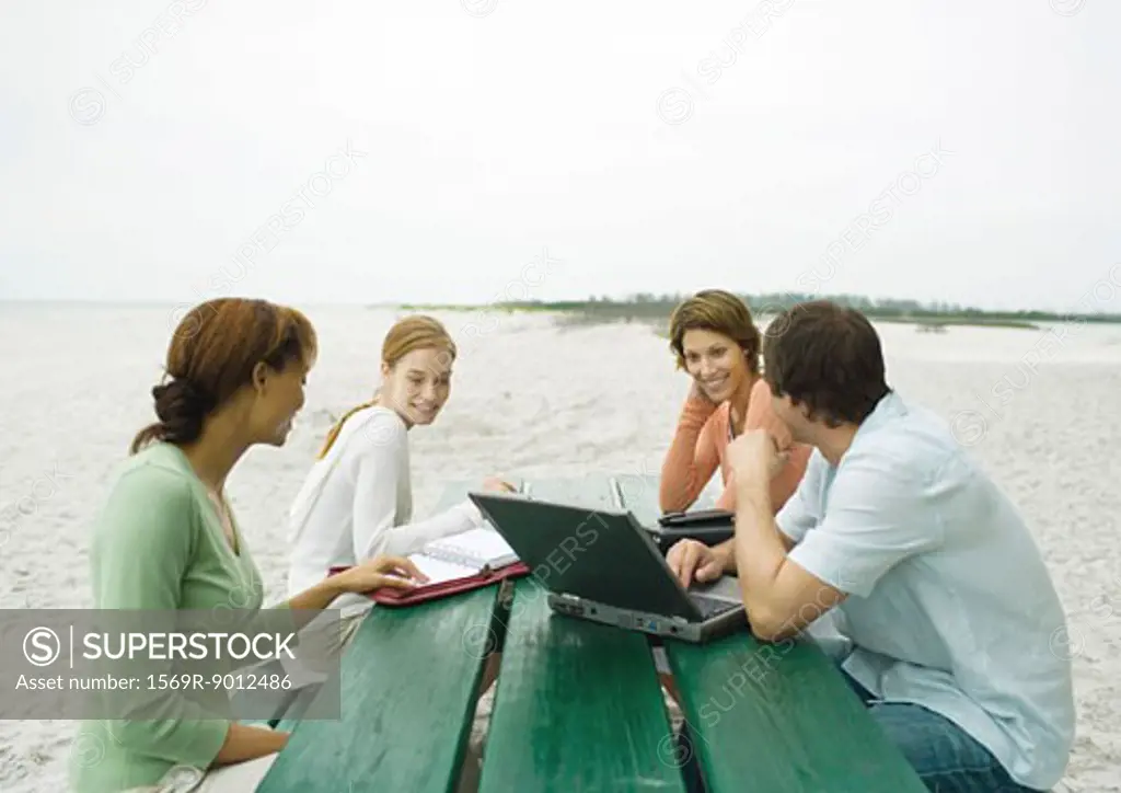 Four casually dressed adults sitting at picnic table on beach, with laptop and agendas