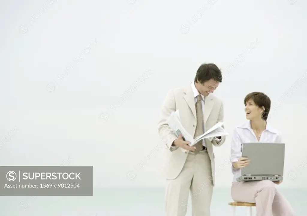 Two business people on beach, one using laptop, the other reading newspaper