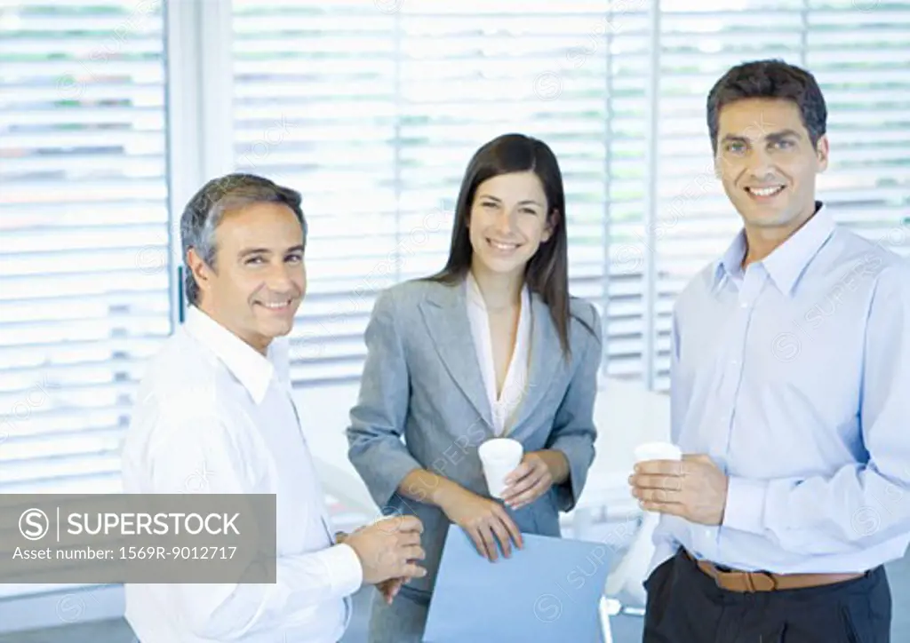 Three business colleagues standing in office with cups in hands, smiling at camera