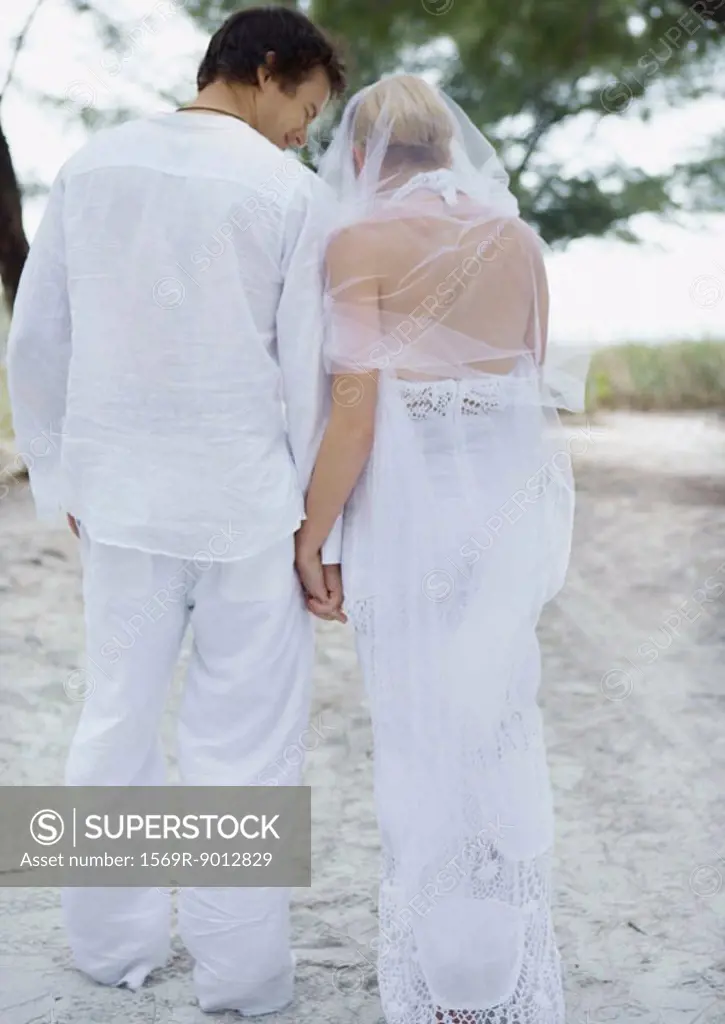 Bride and groom walking on beach, holding hands, rear view