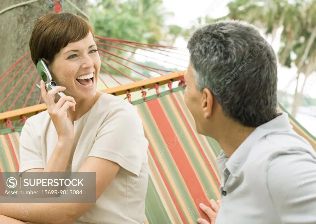Mature couple lounging in hammock, woman using cell phone