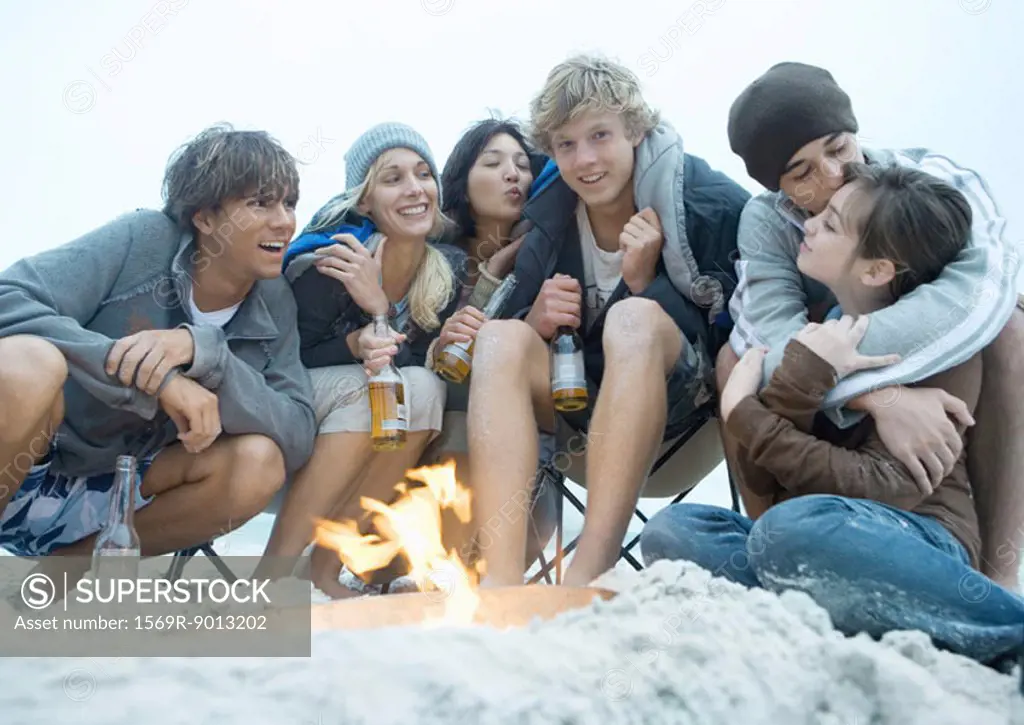 Group of young adults sitting around beach campfire, with bottles of beer