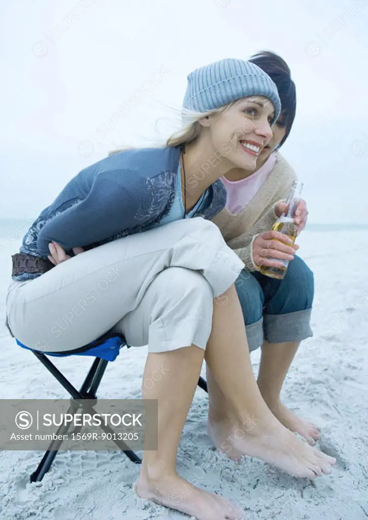 Two young women on beach, leaning forward, on holding beer bottle