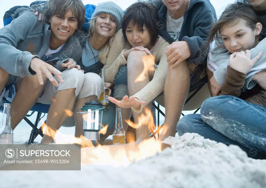 Group of young adult friends, sitting around campfire on beach, reaching hands toward fire