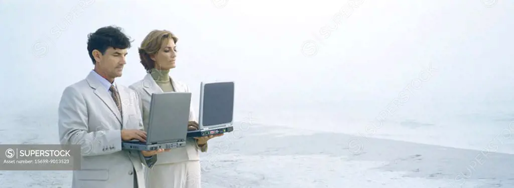 Businessman and woman standing, holding laptops on beach