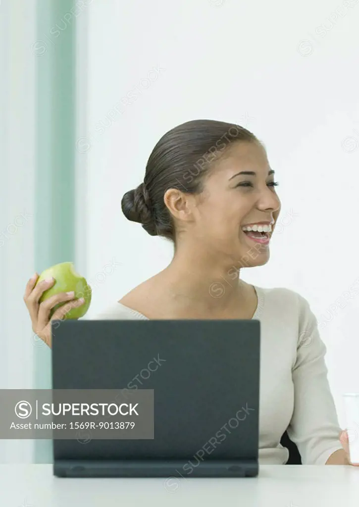 Woman sitting at laptop, holding up apple, laughing