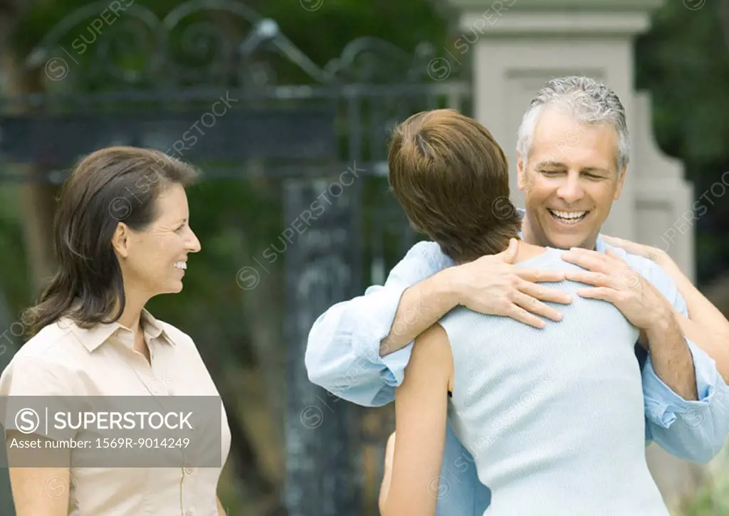 Woman hugging man as second woman watches, smiling