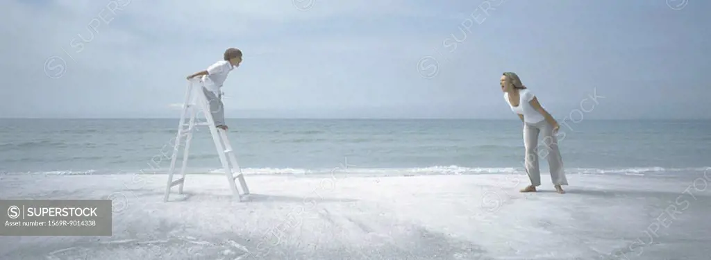 On beach, boy standing on ladder facing woman standing on sand, both screaming